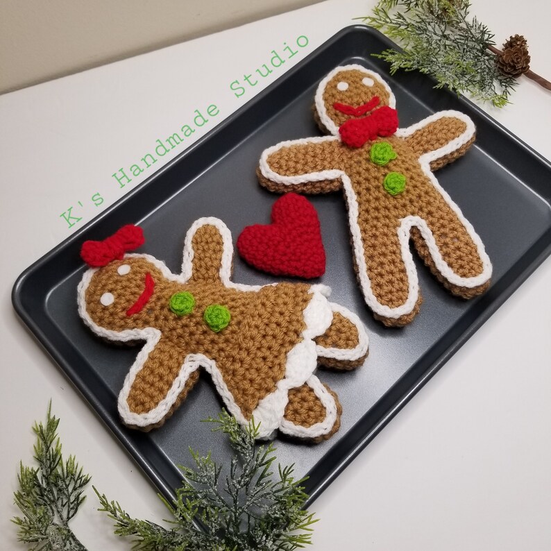 May include: Two crocheted gingerbread people, one with a red bow and the other with a red bow tie, are on a gray baking sheet. A red crocheted heart is between them.