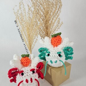 May include: Two white crocheted dragon heads with orange and green accents. The dragon heads are sitting on a brown box. The dragon on the left has red accents and the dragon on the right has green accents. The text "@k.s.handmade.studio" is visible in the background.