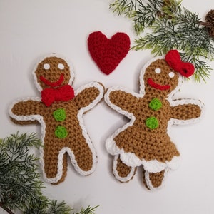 May include: Two crocheted gingerbread people, one with a red bow tie and the other with a red bow, stand next to each other with a red heart between them.