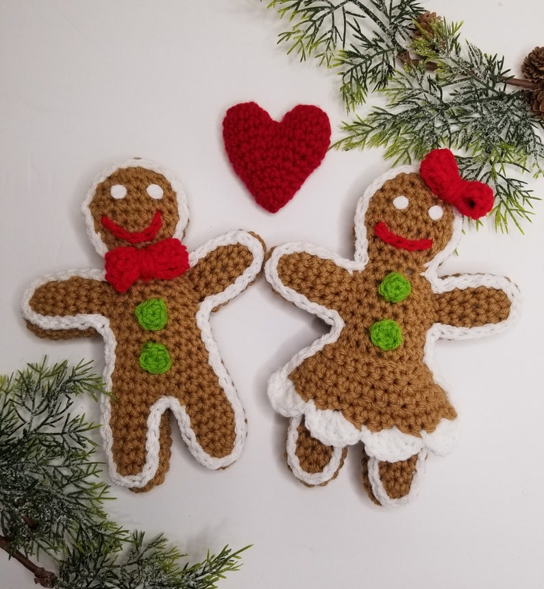 May include: Two crocheted gingerbread people, one with a red bow tie and the other with a red bow, stand next to each other with a red heart between them.
