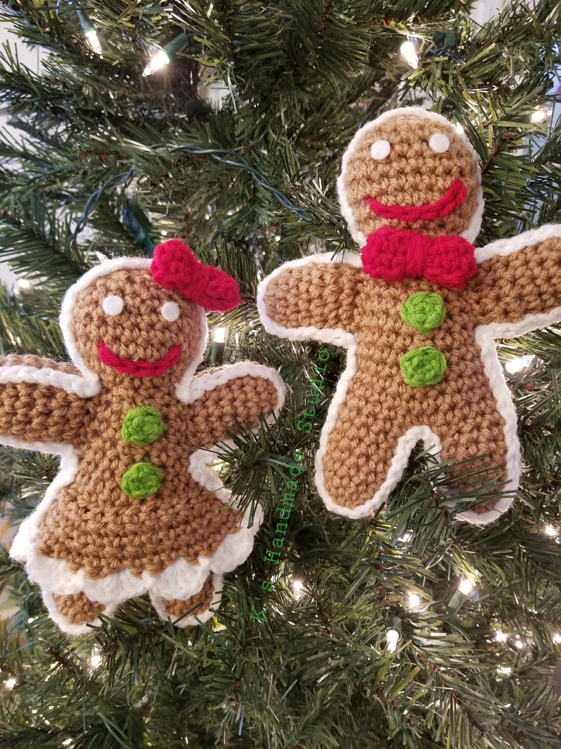 May include: Two crocheted gingerbread people ornaments hanging on a Christmas tree. The gingerbread person on the left is wearing a red bow and the gingerbread person on the right is wearing a red bow tie. Both gingerbread people have green buttons and white icing.