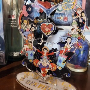 May include: A colorful acrylic stand featuring characters from the anime One Piece. The stand is shaped like a heart with a red heart charm hanging from the top. The stand is on a wooden surface.