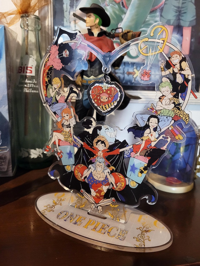 May include: A colorful acrylic stand featuring characters from the anime One Piece. The stand is shaped like a heart with a red heart charm hanging from the top. The stand is on a wooden surface.
