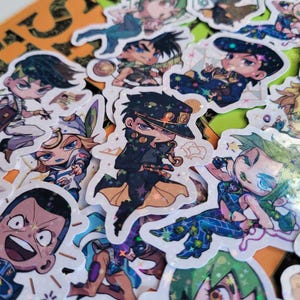 Puede incluir: Una colección de pegatinas de personajes de anime coloridos que presentan diversas poses y expresiones. Las pegatinas están impresas en fondos blancos con acentos de purpurina.