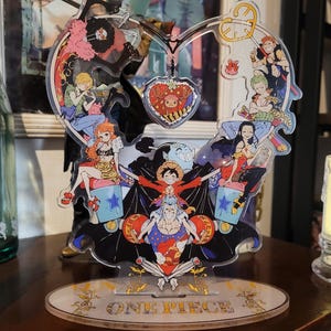 May include: A heart-shaped acrylic stand with a One Piece theme. The stand features characters from the anime series, including Luffy, Zoro, Nami, and Sanji. The stand is decorated with stars, hearts, and other details. The stand is on a wooden surface.