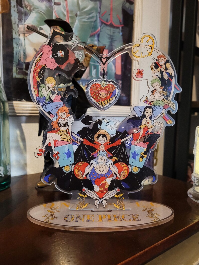 May include: A heart-shaped acrylic stand with a One Piece theme. The stand features characters from the anime series, including Luffy, Zoro, Nami, and Sanji. The stand is decorated with stars, hearts, and other details. The stand is on a wooden surface.