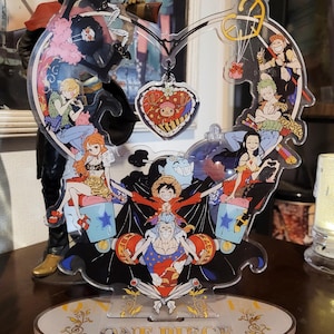 May include: A clear acrylic stand shaped like a heart with a variety of characters from the anime One Piece. The stand features a heart-shaped charm with a character from the show. The stand is on a white oval base with the words "One Piece" printed in gold.