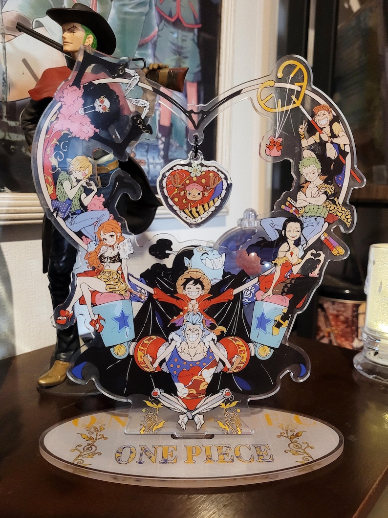 May include: A clear acrylic stand shaped like a heart with a variety of characters from the anime One Piece. The stand features a heart-shaped charm with a character from the show. The stand is on a white oval base with the words "One Piece" printed in gold.