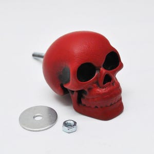 May include: A red skull-shaped object with black shading, a metal rod, a silver washer, and a hex nut. The skull has hollow eye sockets and a detailed jawline. The objects are on a white background.