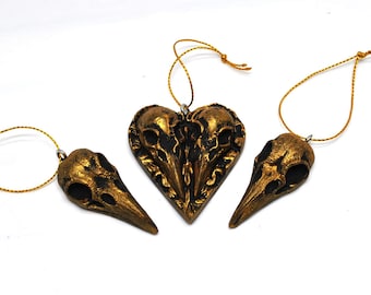 May include: Three raven skull ornaments with gold-colored string for hanging. One ornament is heart-shaped, and the other two are raven skull shaped. The ornaments are painted in a dark gold color with black accents.