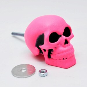 May include: A vibrant pink skull-shaped cabinet knob with black detailing. It features a metal rod, a washer, and a nut. This decorative knob is designed for furniture and adds a unique touch.