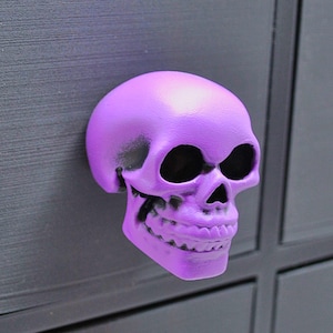 May include: A purple skull-shaped drawer pull. The skull has black eye sockets and a toothy grin.