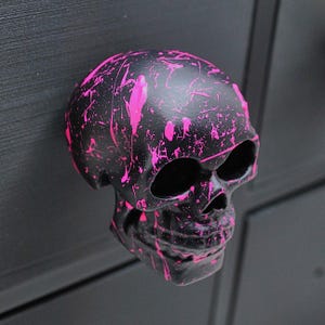 May include: Black skull-shaped decorative object with bright pink paint splatter design. The skull has hollow eye sockets and a detailed facial structure. The object is likely made of a hard material and is mounted on a dark surface.