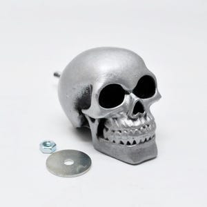May include: A silver-colored skull-shaped object with a detailed design, featuring eye sockets, a nose cavity, and teeth. A metal washer and nut are next to the skull. The skull is likely a decorative item or part of a larger assembly.