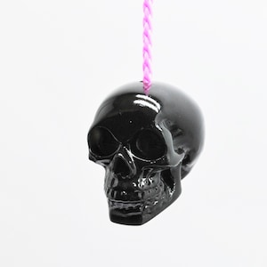 May include: A black skull pendant with a shiny finish hangs from a pink cord.