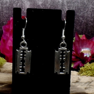 May include: A pair of silver earrings shaped like razor blades, displayed on a black velvet stand.