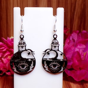 May include: A pair of black dangle earrings with a silver hook. The earrings are shaped like a bottle with a crescent moon, stars, and an eye design.