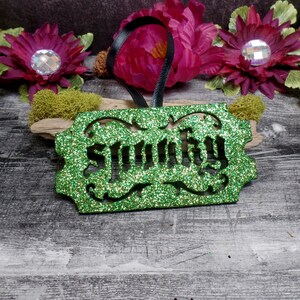 Puede incluir: Adorno verde brillante con la palabra "Sunny" recortada en el centro. El adorno tiene un borde decorativo y una cinta negra para colgar. El fondo incluye flores y musgo.