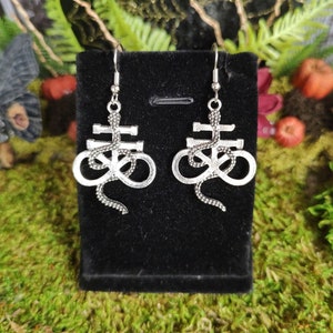 May include: A pair of silver earrings with a snake wrapped around a pentagram symbol. The earrings are displayed on a black velvet backing.