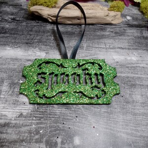 Puede incluir: Un adorno verde brillante con la palabra "Spooky" recortada en una fuente decorativa. El adorno tiene una cinta negra para colgar. El fondo es una superficie de madera gris.