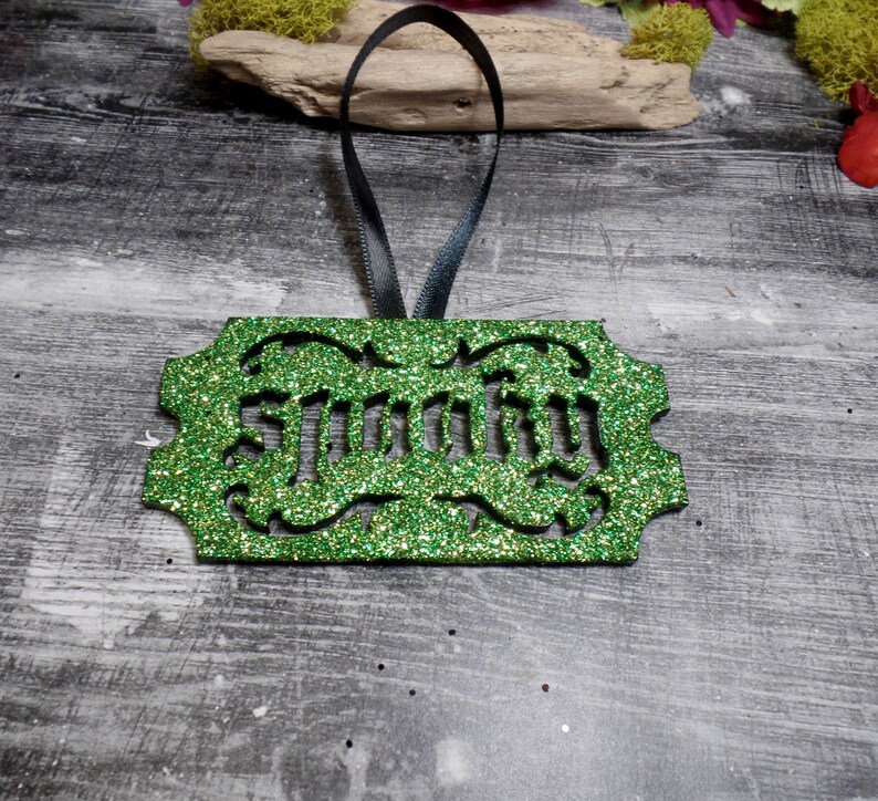 Puede incluir: Un adorno verde brillante con la palabra "Spooky" recortada en una fuente decorativa. El adorno tiene una cinta negra para colgar. El fondo es una superficie de madera gris.