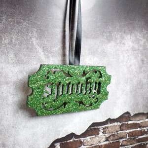 Puede incluir: Un adorno verde brillante con la palabra "Murphy" recortada. El adorno rectangular tiene bordes decorativos y cuelga de una cinta negra. El fondo es una superficie gris texturizada con una pared de ladrillos.