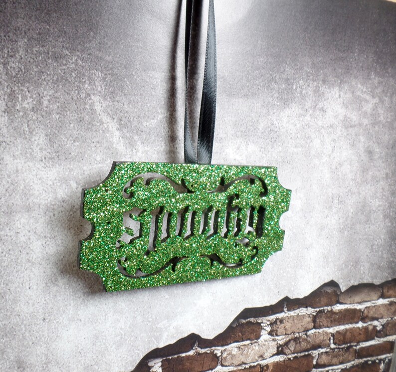Puede incluir: Un adorno verde brillante con la palabra "Murphy" recortada. El adorno rectangular tiene bordes decorativos y cuelga de una cinta negra. El fondo es una superficie gris texturizada con una pared de ladrillos.