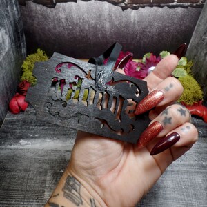 Puede incluir: Una etiqueta decorativa de madera negra con la palabra "Witch" recortada en una fuente g&oacute;tica. La etiqueta tiene remolinos decorativos y est&aacute; sujeta a una cinta negra. El fondo incluye flores y musgo.