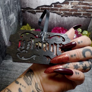 May include: A dark wooden ornament with the word "WITCH" cut out. The ornament has a black ribbon and a silver charm. The background includes a brick wall and floral accents. The ornament is held in a hand with long, dark red nails.