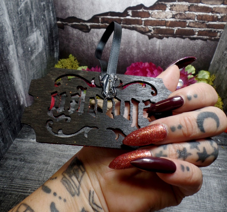 May include: A dark wooden ornament with the word "WITCH" cut out. The ornament has a black ribbon and a silver charm. The background includes a brick wall and floral accents. The ornament is held in a hand with long, dark red nails.