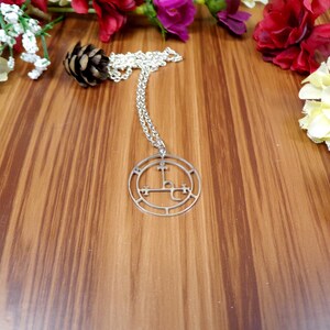 May include: Silver necklace with a circular pendant featuring a sigil design. The pendant is on a wooden surface with flowers.