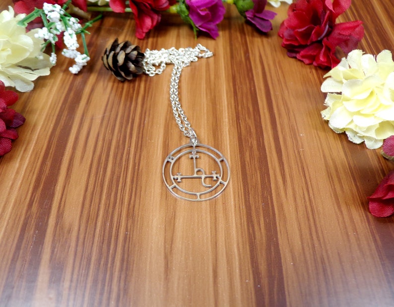 May include: Silver necklace with a circular pendant featuring a sigil design. The pendant is on a wooden surface with flowers.