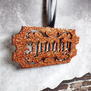 May include: An orange glitter ornament with the word "Sunny" cut out. The ornament has a decorative border and is suspended by a grey ribbon. The background is a textured grey surface.