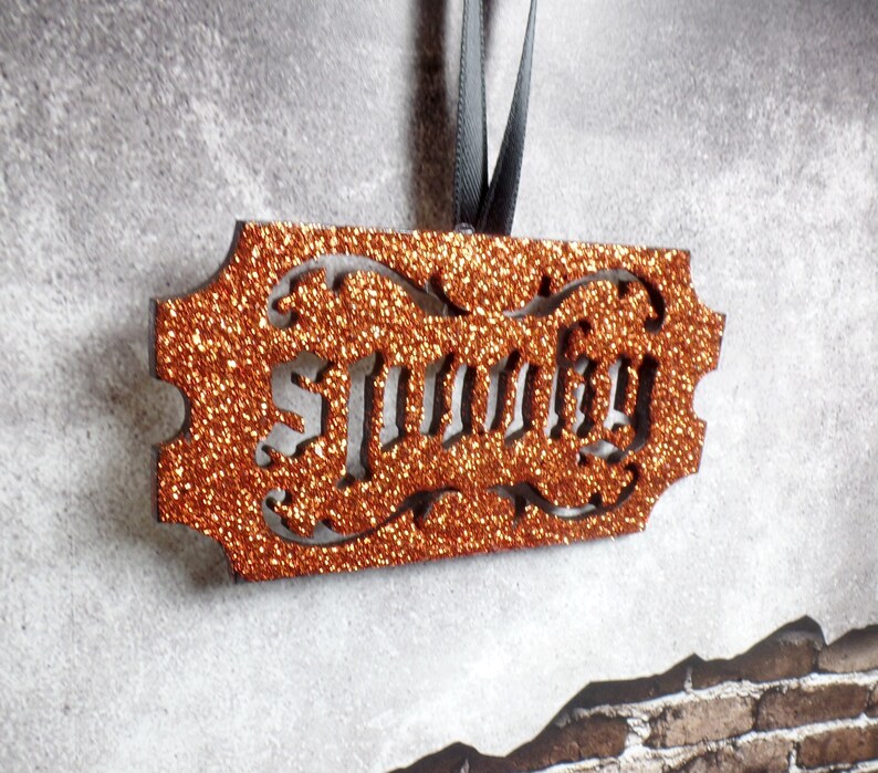 May include: An orange glitter ornament with the word "Sunny" cut out. The ornament has a decorative border and is suspended by a grey ribbon. The background is a textured grey surface.