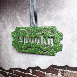 Puede incluir: Adorno verde brillante con la palabra "Spooky" recortada. El adorno rectangular tiene bordes decorativos y est&aacute; suspendido por una cinta gris. El fondo es una pared gris texturizada con un patr&oacute;n de ladrillos.