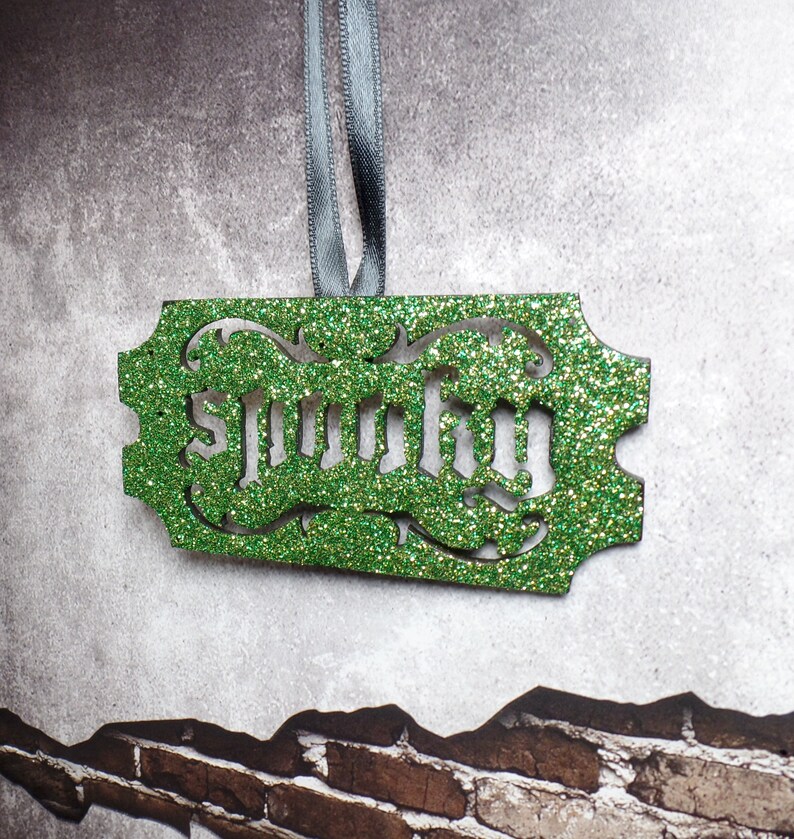 Puede incluir: Adorno verde brillante con la palabra "Spooky" recortada. El adorno rectangular tiene bordes decorativos y est&aacute; suspendido por una cinta gris. El fondo es una pared gris texturizada con un patr&oacute;n de ladrillos.