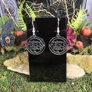 May include: Silver-colored dangle earrings with a circular design featuring an intricate, stylized symbol. The earrings are displayed on a black stand against a backdrop of greenery, small pumpkins, and a spiderweb design.