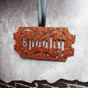 May include: An orange glitter-covered rectangular ornament with the word "Spooky" cut out in a gothic font. It hangs from a grey ribbon. The background is a textured grey wall.