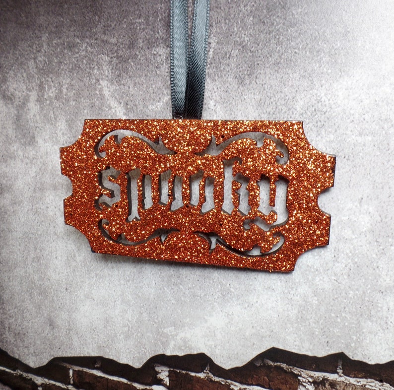May include: An orange glitter-covered rectangular ornament with the word "Spooky" cut out in a gothic font. It hangs from a grey ribbon. The background is a textured grey wall.