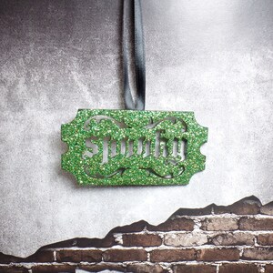 Puede incluir: Un adorno verde brillante con el nombre "Smitty" recortado en una fuente decorativa. El adorno rectangular tiene bordes ornamentados y cuelga de una cinta gris. El fondo es una superficie gris texturizada con un patr&oacute;n de ladrillos.