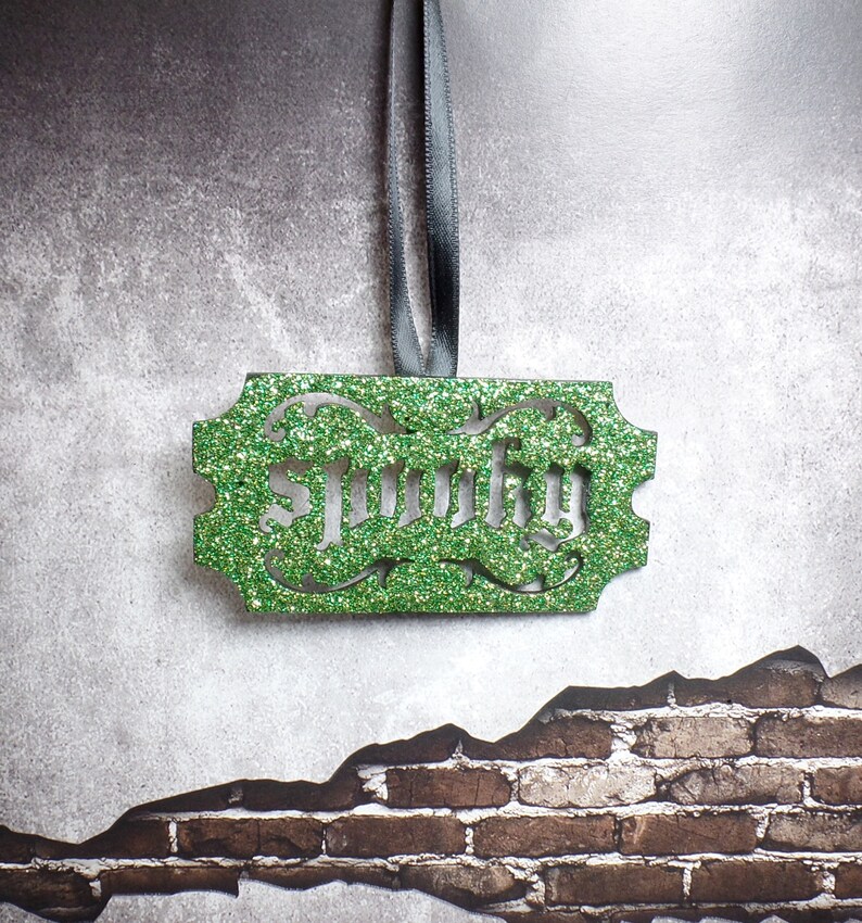 Puede incluir: Un adorno verde brillante con el nombre "Smitty" recortado en una fuente decorativa. El adorno rectangular tiene bordes ornamentados y cuelga de una cinta gris. El fondo es una superficie gris texturizada con un patr&oacute;n de ladrillos.