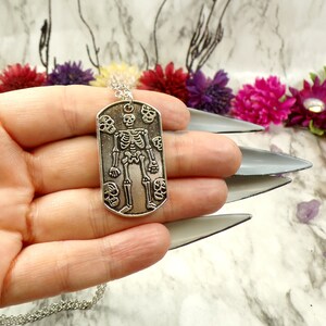 May include: Silver skeleton pendant necklace with a dog tag style charm featuring a skeleton with skulls around it.