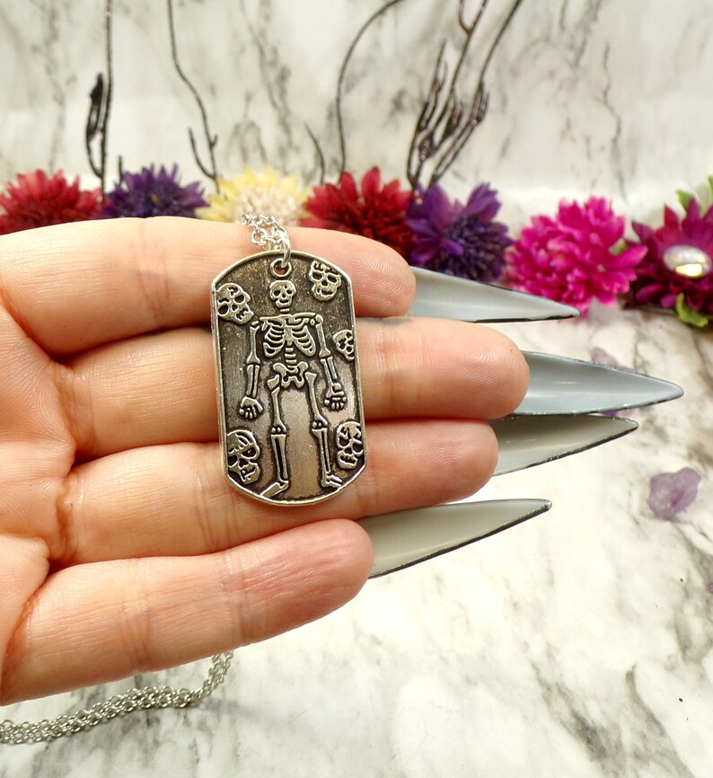 May include: Silver skeleton pendant necklace with a dog tag style charm featuring a skeleton with skulls around it.
