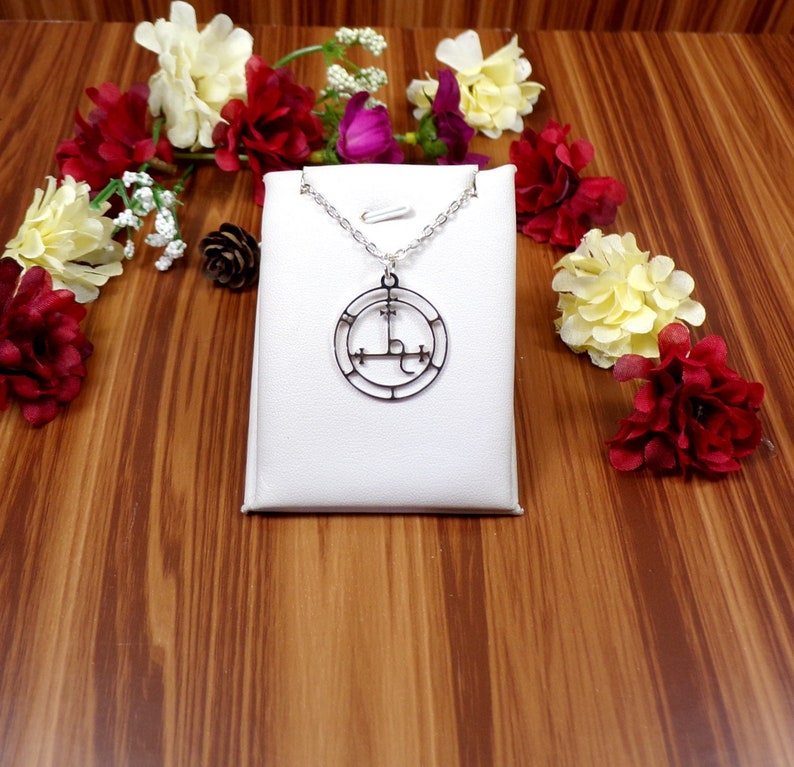 May include: A silver necklace with a circular pendant featuring a black sigil. The pendant is on a white display stand against a wood background.
