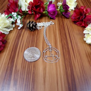 May include: A silver necklace with a circular pendant featuring a sigil design. The pendant is surrounded by a circle with a series of symbols. The necklace is displayed on a wooden surface with artificial flowers.