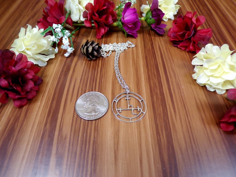 May include: A silver necklace with a circular pendant featuring a sigil design. The pendant is surrounded by a circle with a series of symbols. The necklace is displayed on a wooden surface with artificial flowers.