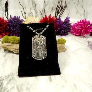 May include: Silver skeleton pendant necklace with a dog tag style charm featuring a skeleton surrounded by skulls. The necklace is displayed on a black velvet pouch.