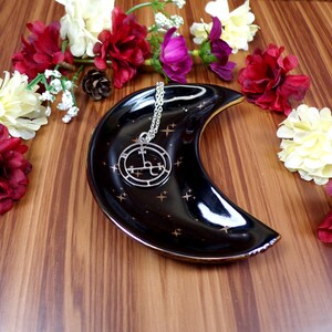 May include: A silver necklace with a black and silver pendant featuring a sigil design. The necklace is displayed on a black crescent moon-shaped dish with gold stars.