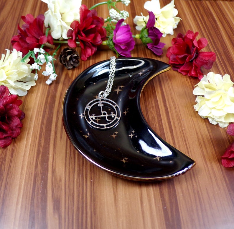 May include: A silver necklace with a black and silver pendant featuring a sigil design. The necklace is displayed on a black crescent moon-shaped dish with gold stars.