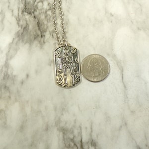 May include: Silver necklace with a skeleton pendant surrounded by skulls. The pendant is shaped like a dog tag.